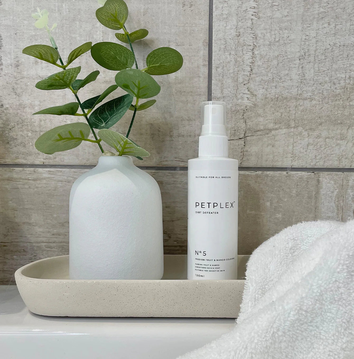 White bottle of dog cologne labelled 'PETPLEX' on a tray with a vase and plant against a tiled wall.
