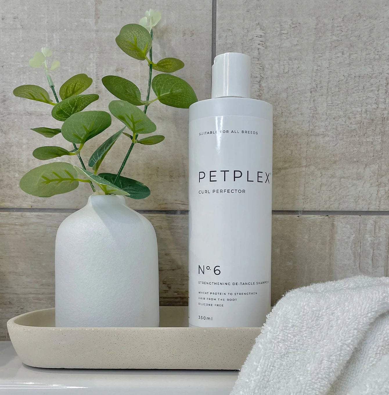PETPLEX curly coat dog shampoo bottle on a tray with a vase and plant against a tiled wall.