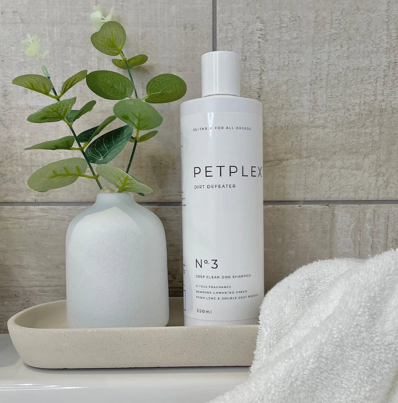 PETPLEX dirt defeater deep clean dog shampoo bottle on a tray with a vase and plant against a tiled wall.