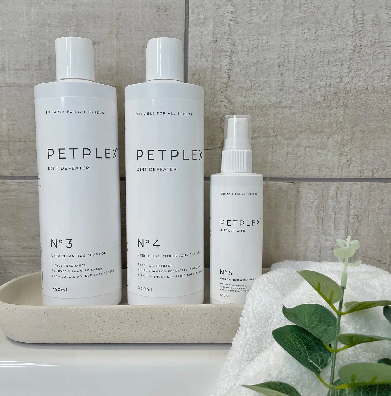 Three Petplex pet care products, including dirt defeater deep clean dog shampoo, dog conditioner, and dog cologne, all on a tray together with a towel and plant in the background.