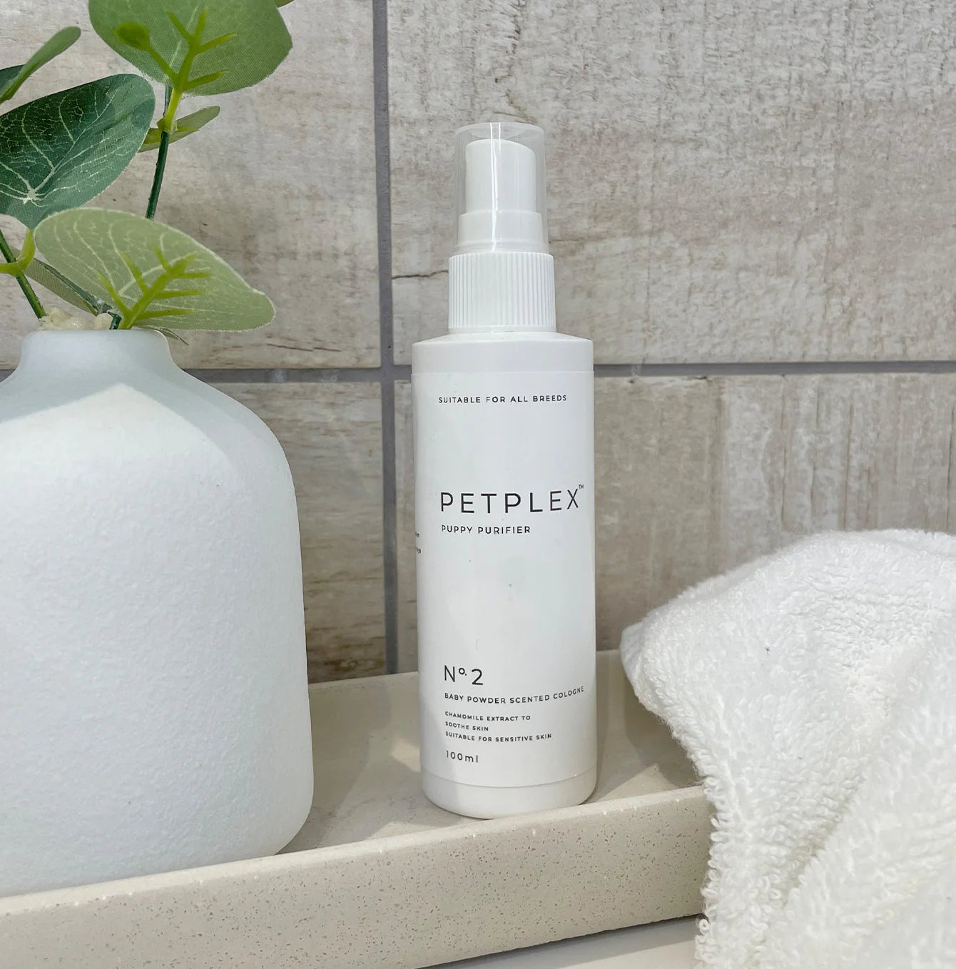 White bottle of PETPLEX puppy cologne on a shelf with a towel and plant in the background