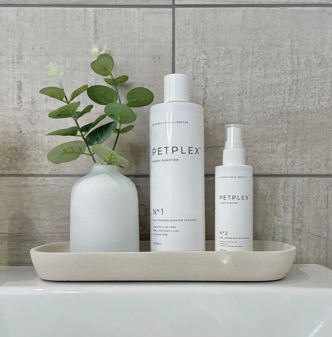 Two bottles of PETPLEX puppy shampoo and cologne on a tray with a white vase and green plant against a tiled wall.